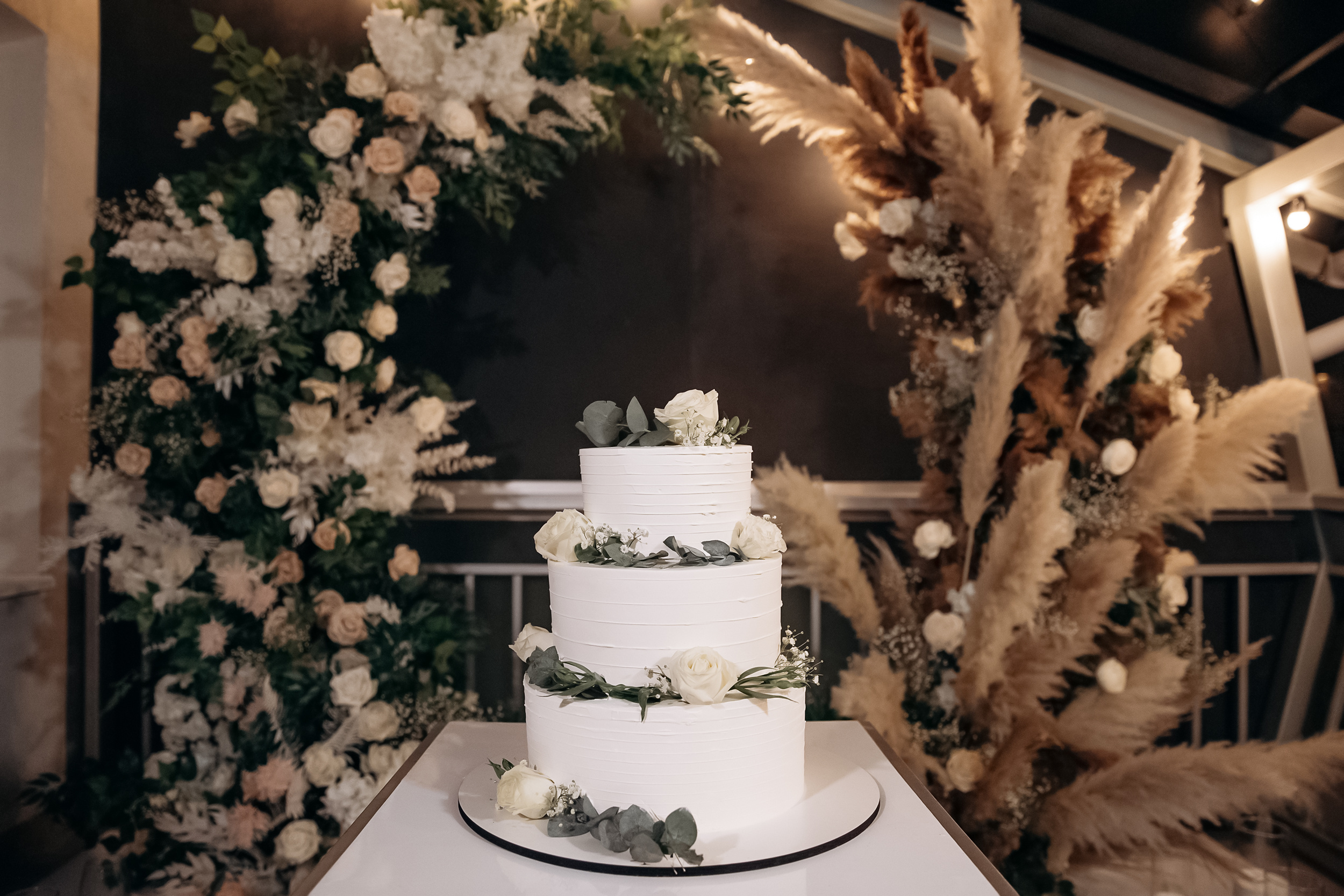 Large luxury wedding cake on a background of a flowering arch in the style of boho. Wedding dessert under the light of evening light bulbs. Wedding decor.